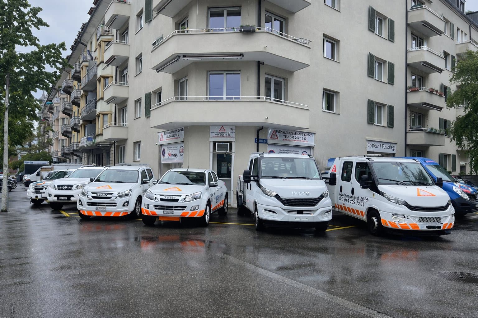 Mehrere weisse Einsatzfahrzeuge der Abschleppdienst Zürich GmbH parkiert auf einer nassen Strasse vor einem Wohnhaus.